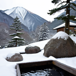 **A serene open-air onsen (rotenburo) nestled amidst a snowy mountain landscape in the Japanese Alps. The warm, steaming water contrasts with the cold, crisp air. Incorporate natural elements like large boulders and evergreen trees. Focus on capturing the sense of tranquility and harmony with nature.**